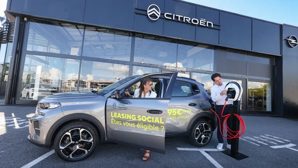 Une femme prend possession de sa citroen acquise via le leasing social devant une concession citroen avec un commercial qui lui explique le fonctionnement d'une borne de recharge.