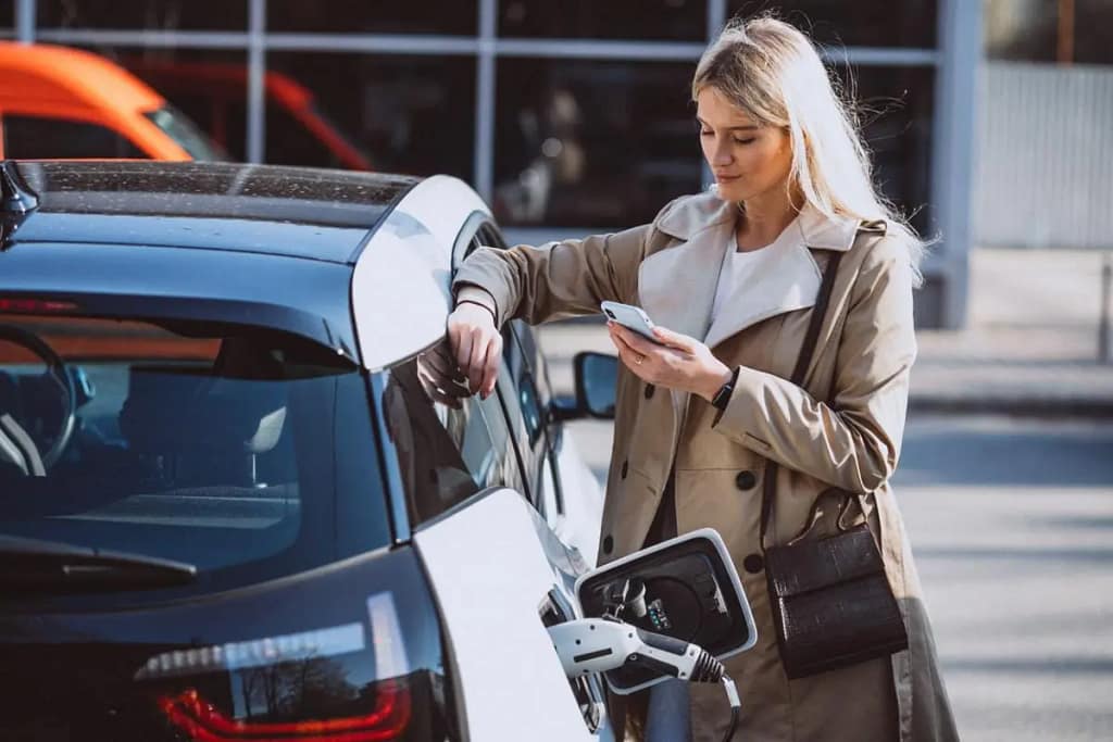 Une femme sur un parking est appuyé sur sa voiture et consulte son téléphone portable